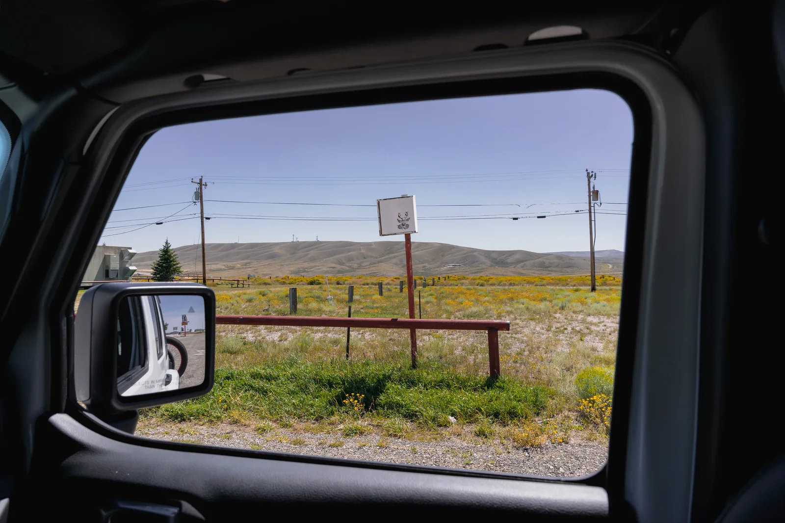 View from inside a parked vehicle through the open passenger window, with a wing mirror, a wooden rail, dry grassland and bare hills under a clear sky in strong sun