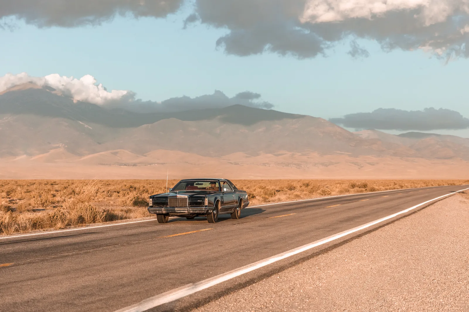 A dark vintage car driving toward the left on an empty desert highway, placed left of centre, with the road stretching away behind it to the right beneath distant mountains