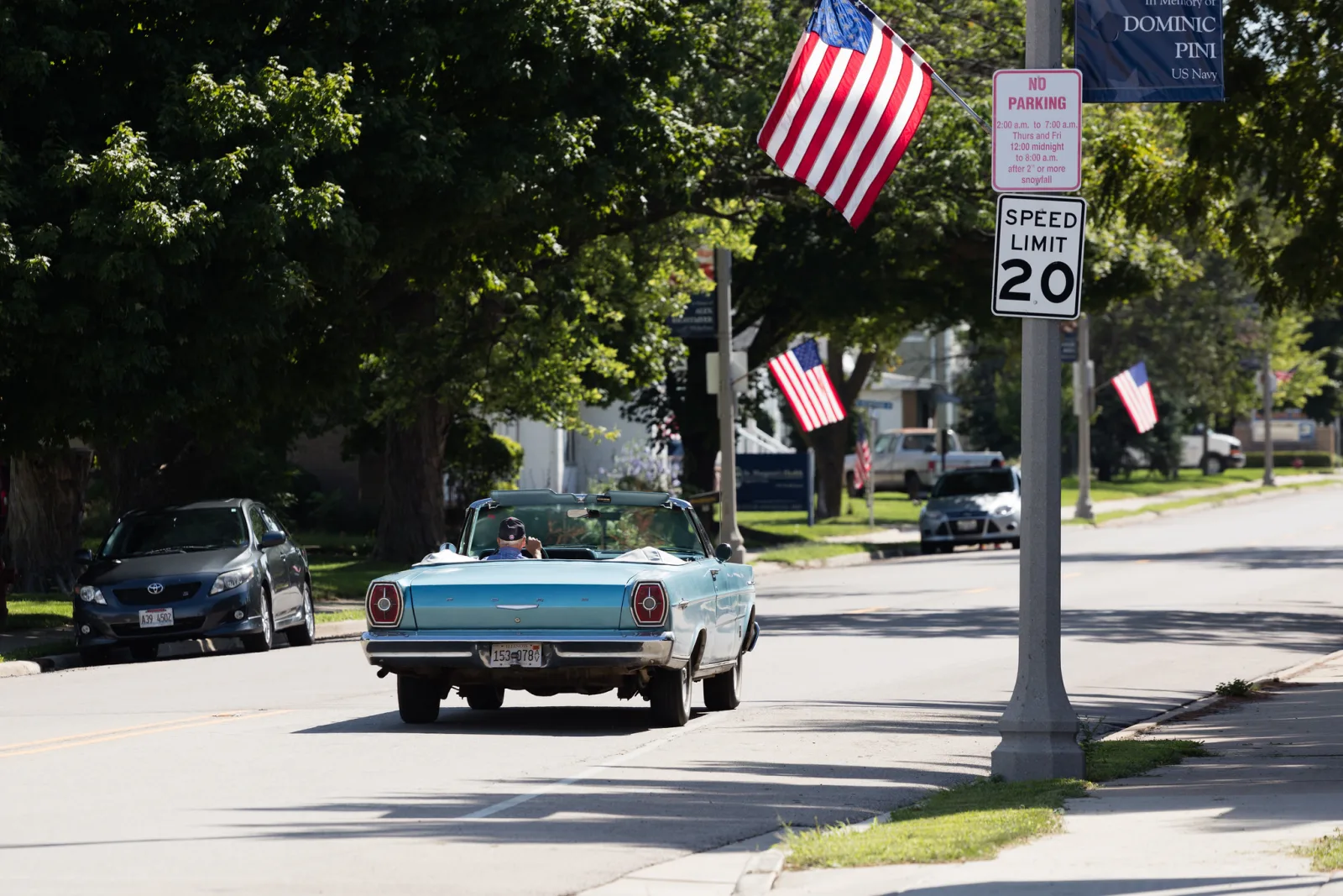 A light blue vintage convertible driving down a tree-lined small town street with American flags on the lamp posts and a modern car parked behind it