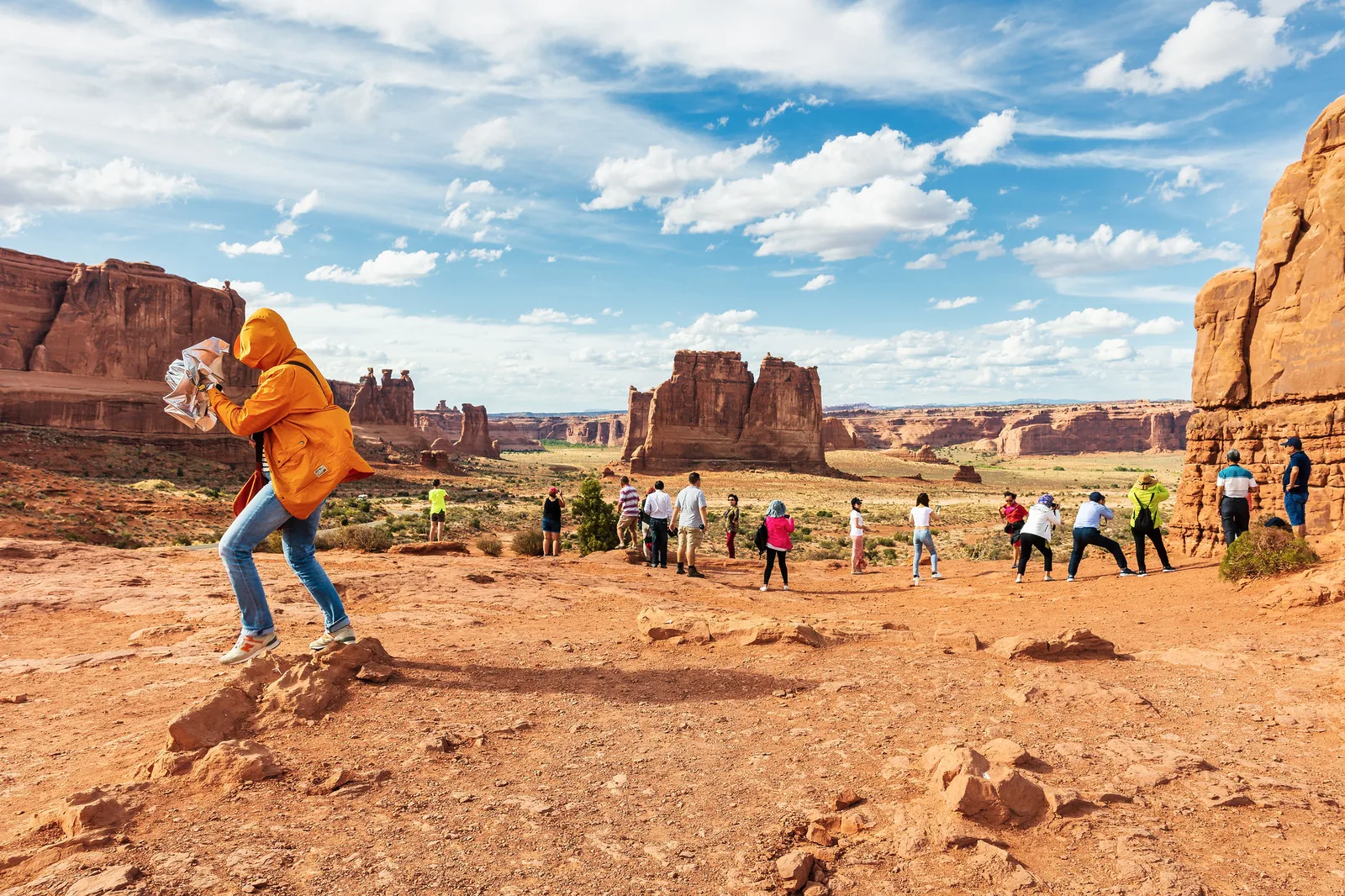 A person in an orange jacket crouching to photograph a line of visitors standing on open red desert ground, with sandstone buttes and a cloud-filled blue sky behind them