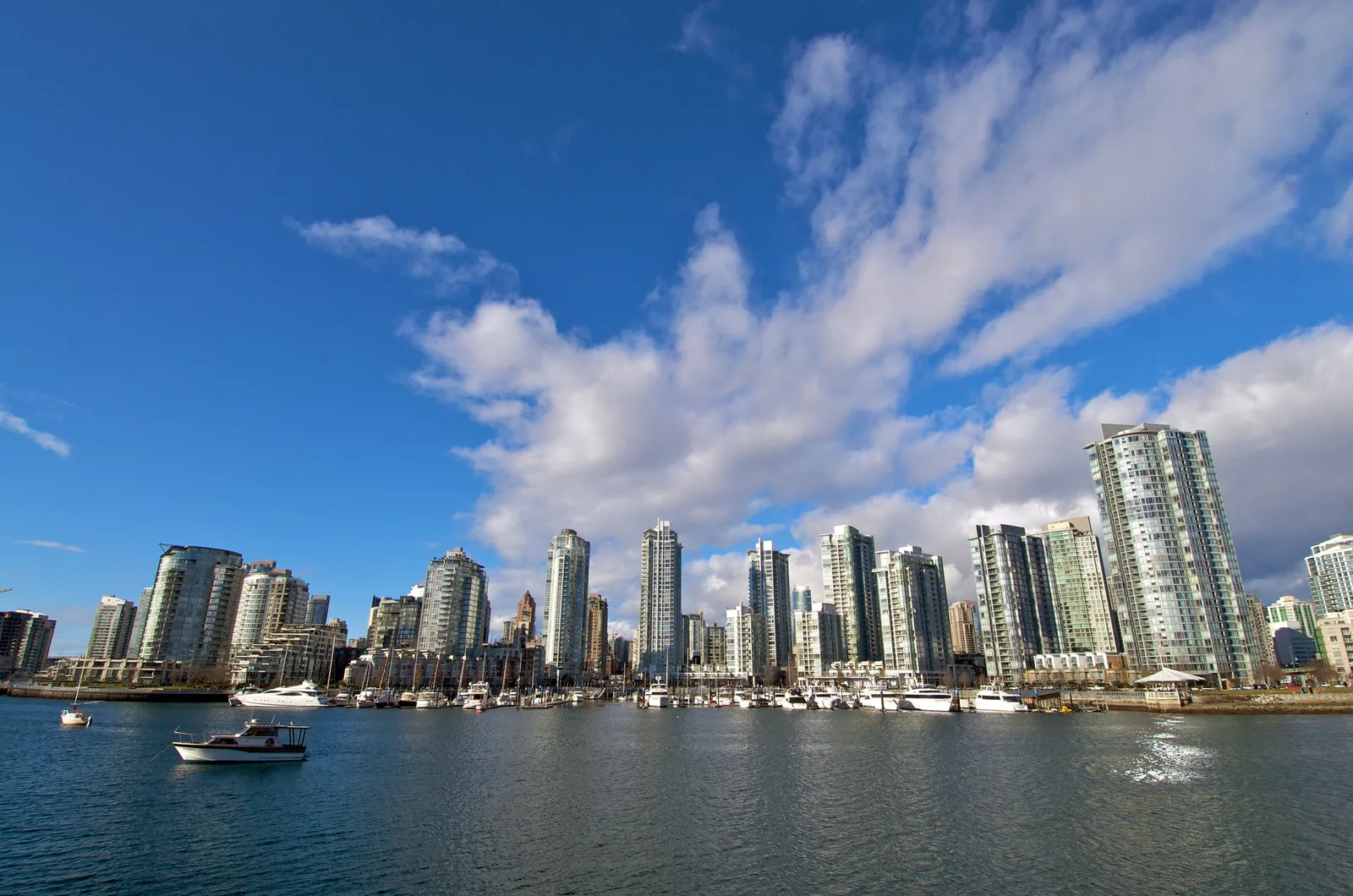 A row of glass residential towers along a marina under a deep blue sky with large white clouds, boats moored on the water