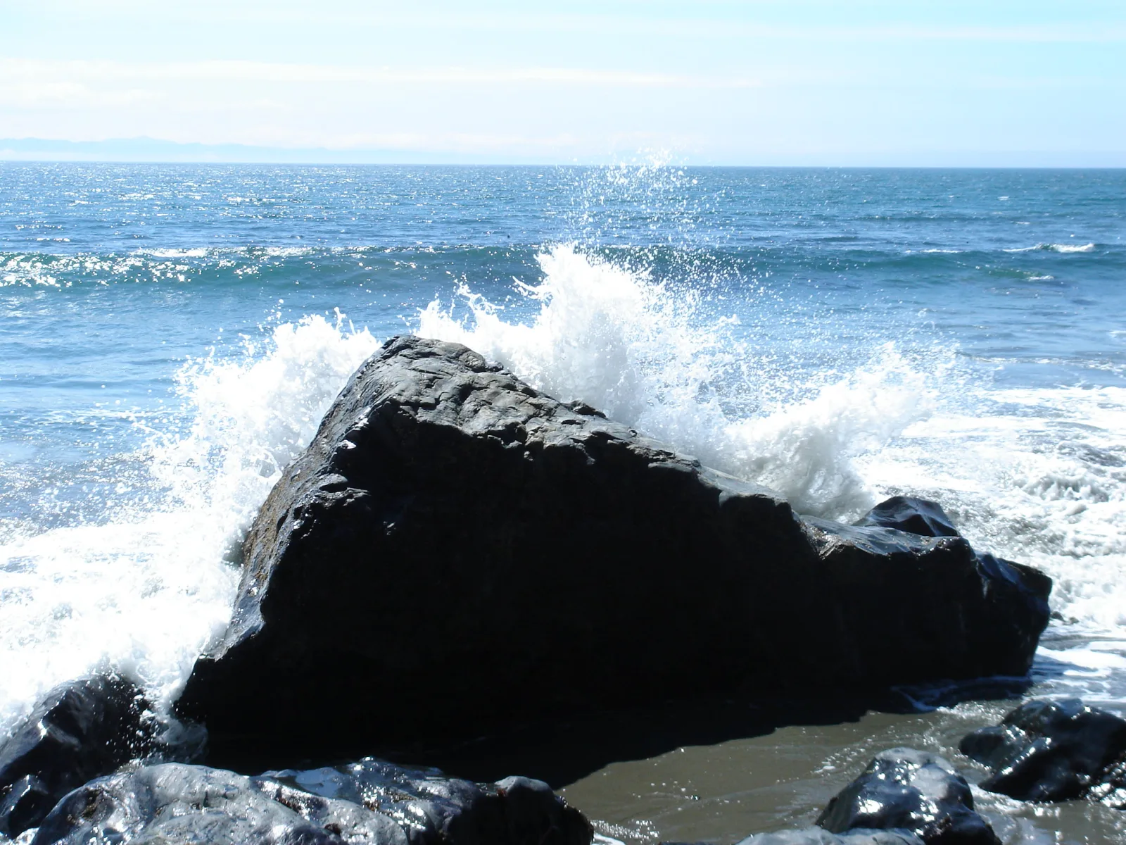 A wave breaks against a large dark rock on a sunny shore, throwing white spray into the air above it