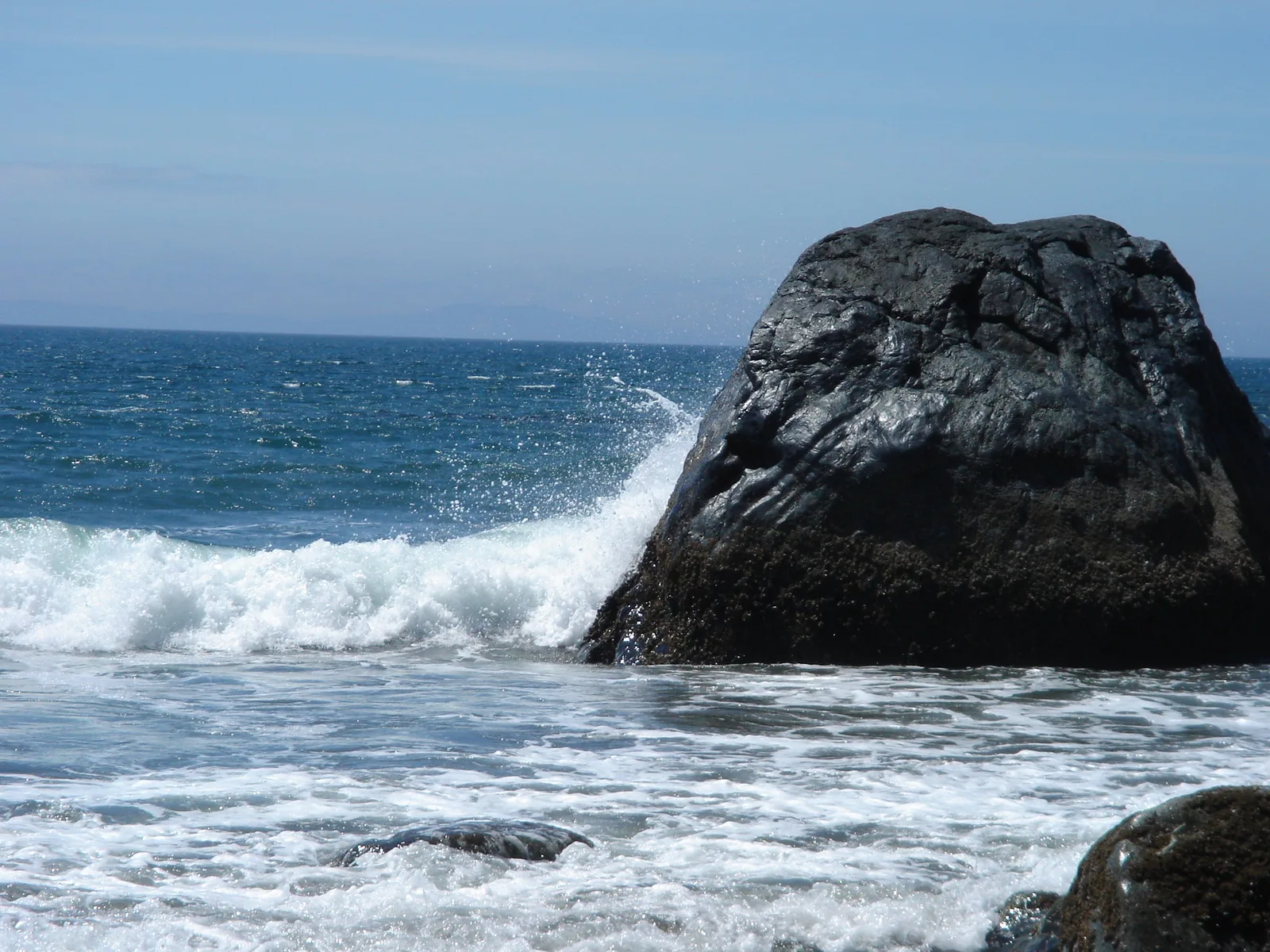 A wave breaking over a large dark boulder on a rocky shore, with the sea horizon behind it sloping noticeably to one side