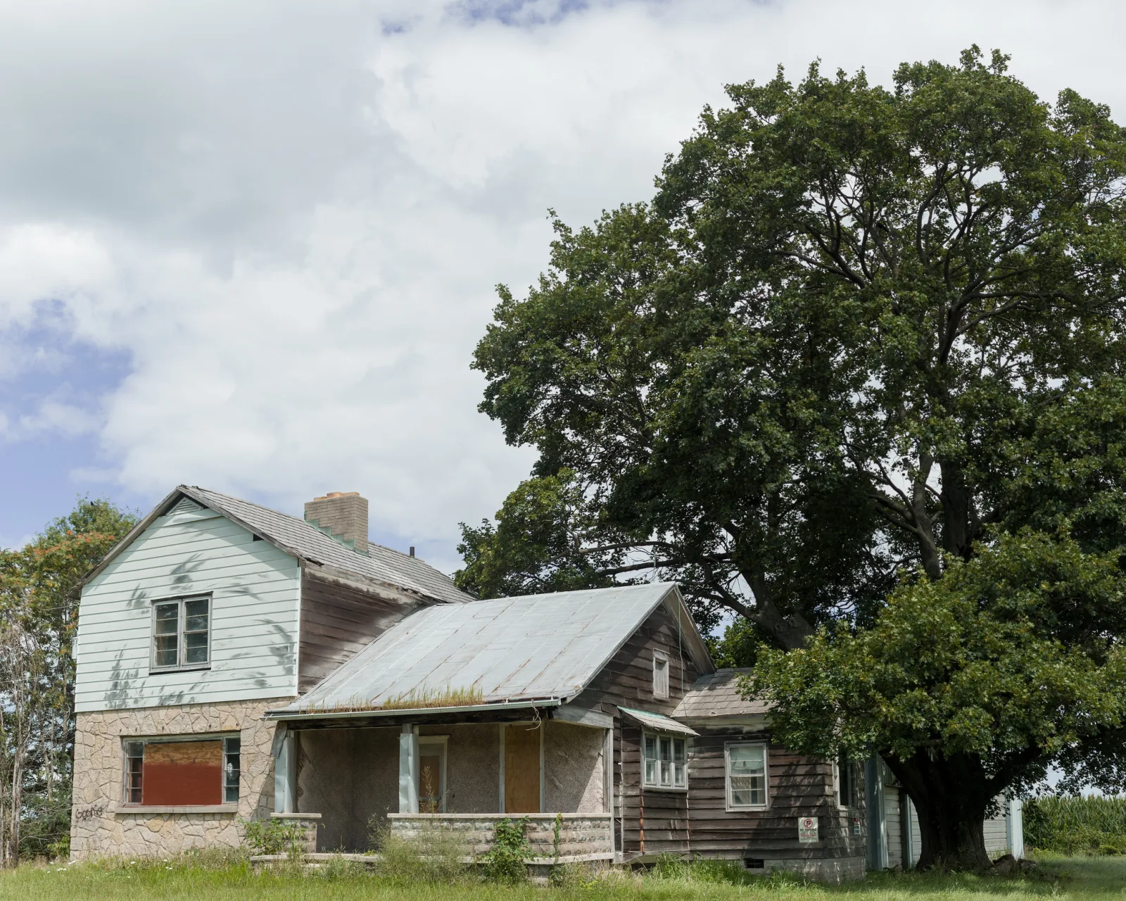 A weathered, abandoned two-storey wooden house with broken windows and sagging porch on a grassy prairie at dusk