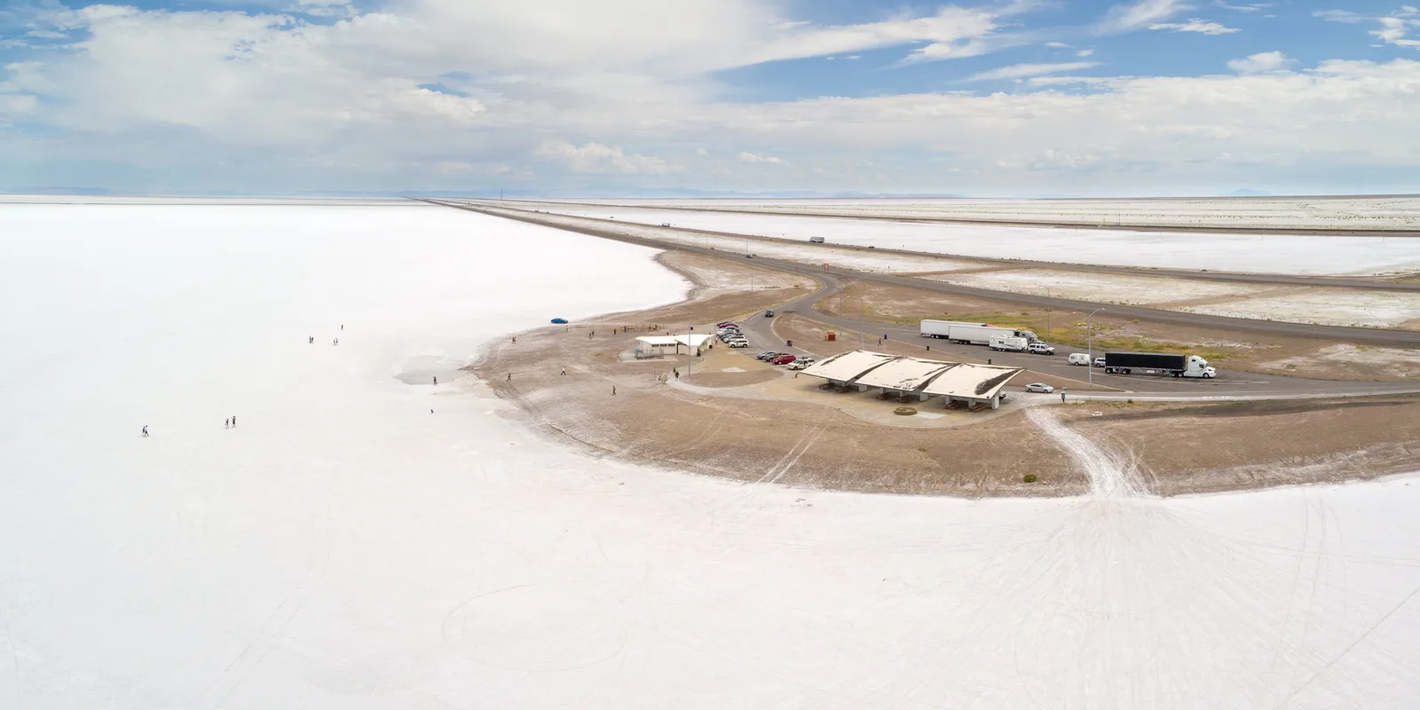 Aerial view of a brilliant white salt flat stretching to the horizon, with a road, a rest area and parked trucks on a strip of brown ground under a partly cloudy sky