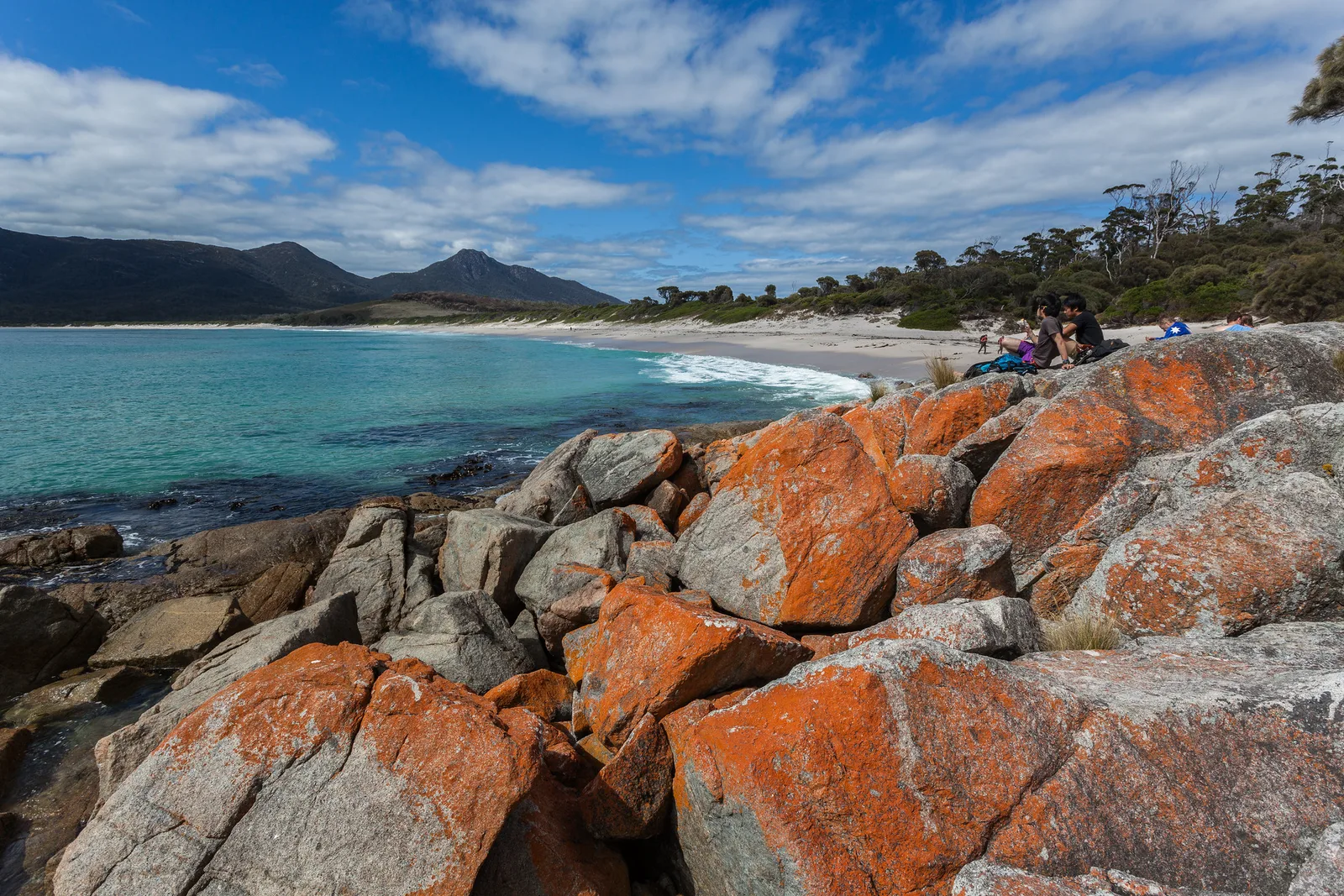 Large granite boulders covered in orange lichen filling the lower half of the frame, with a turquoise bay, a white beach and dark hills small in the distance under a blue sky