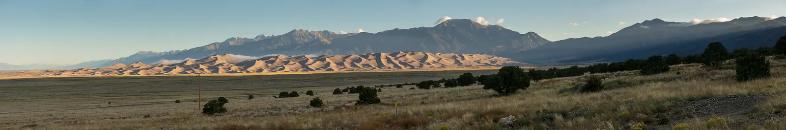 A very wide panorama of pale sand dunes below a mountain range, with dry grassland in the foreground under a clear dawn sky