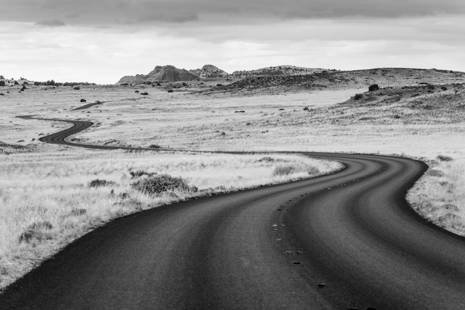 A black and white landscape of a dark gravel road winding through pale grassland toward low hills under a cloudy sky
