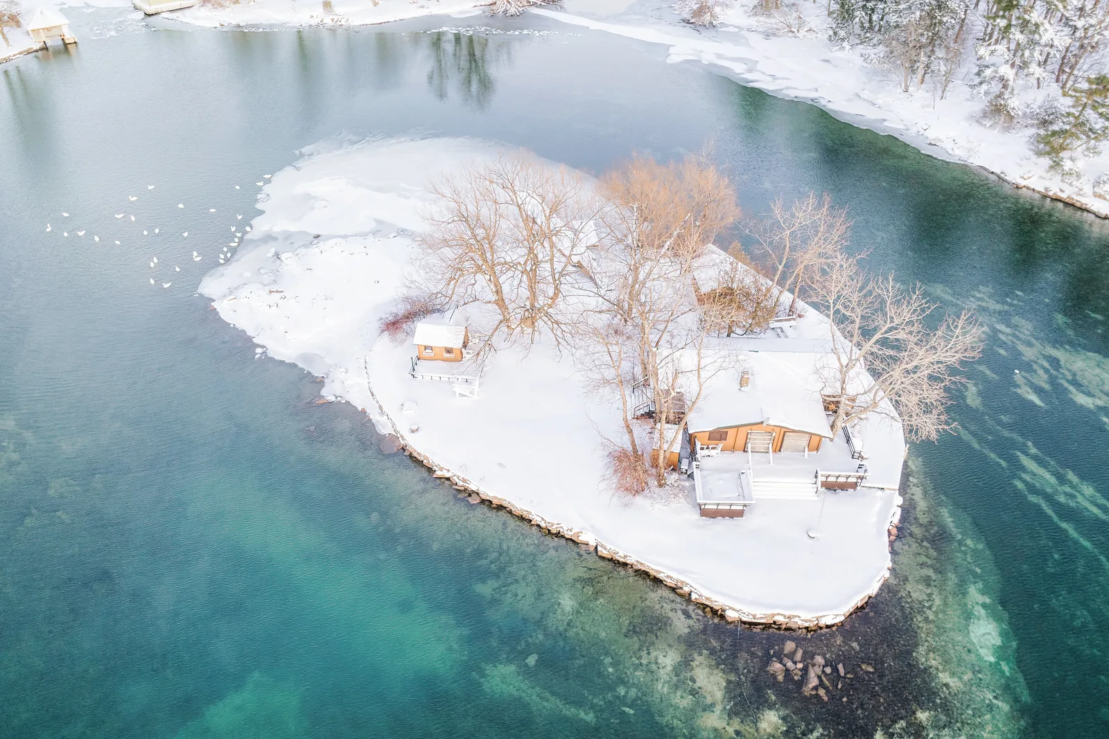 Aerial view of a small snow-covered island with bare trees and a cabin, surrounded by turquoise water and ice