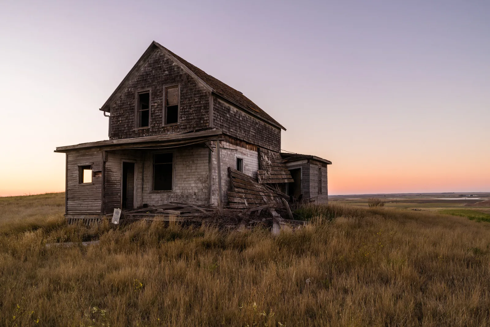 An abandoned two-story wooden farmhouse with a sagging lean-to stands in tall dry grass under a pink and orange dusk sky.