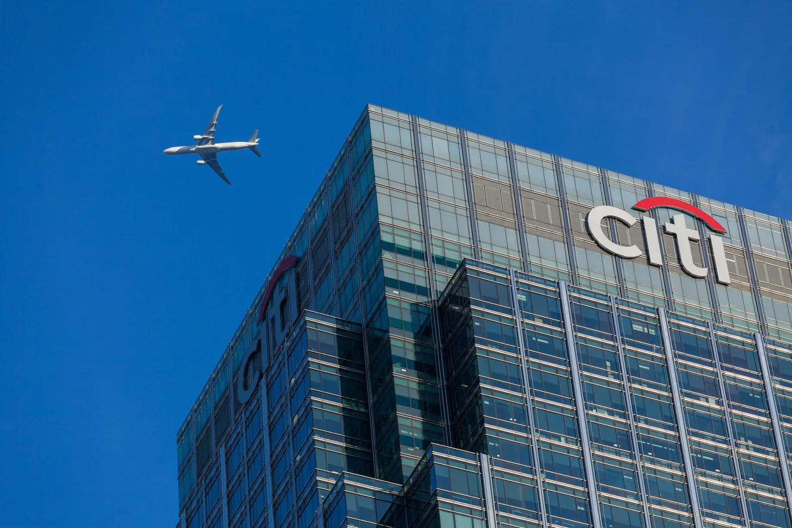 An airliner crosses a clear blue sky above a glass office tower.