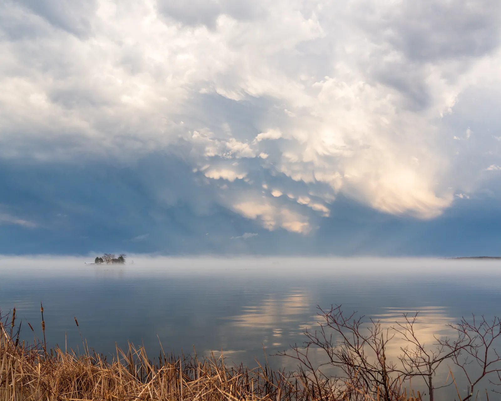 Heavy, dramatic storm clouds over calm water with a small boat on the horizon and dry reeds in the foreground