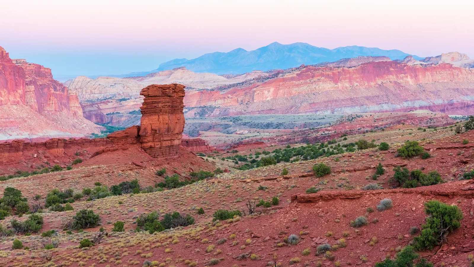 Soft pastel pink and blue sky over red rock cliffs and a lone butte, a gentle gradient like a painted backdrop