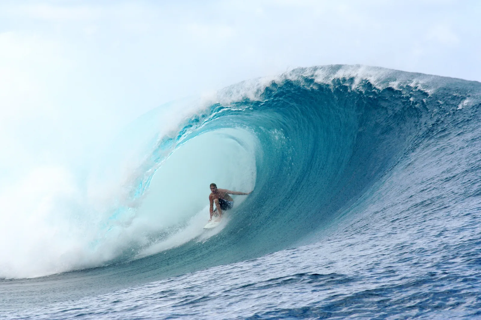 A surfer rides deep inside the curling blue tube of a huge breaking wave, with white spray along its lip.