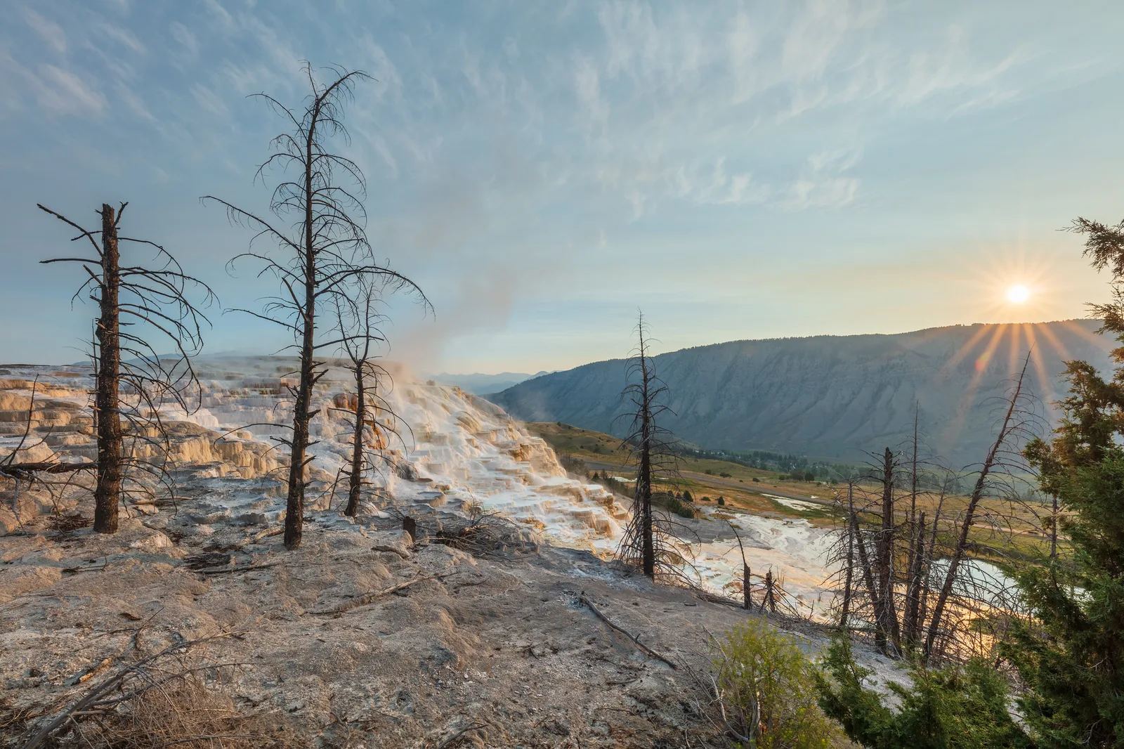 Sunburst rising over a mountain ridge beside steaming white mineral terraces and bare dead trees