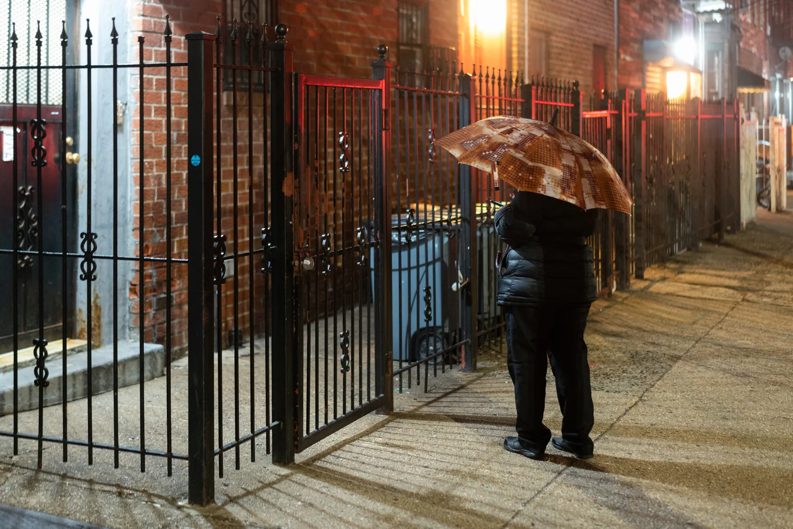 A person seen from behind under a patterned umbrella on a lamp-lit city sidewalk at night, face hidden