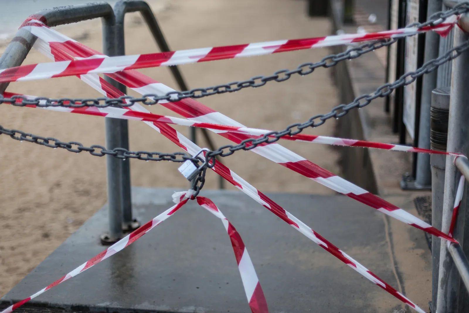 Red and white warning tape and grey chains with a padlock stretched across a closed walkway leading down to a sandy beach