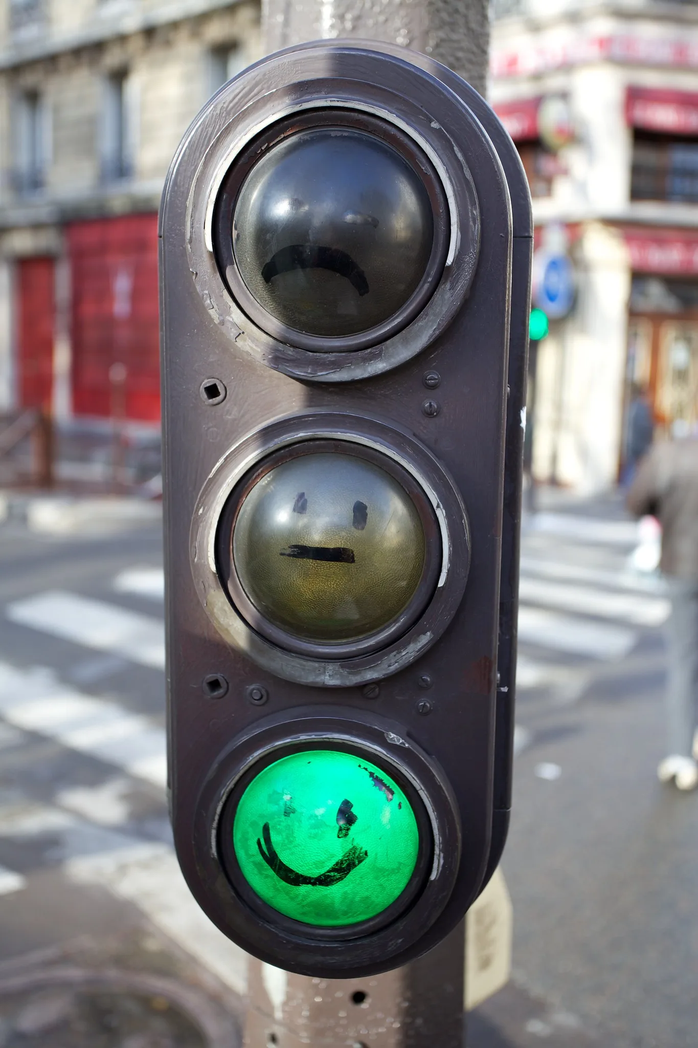 A street traffic light with frowning, neutral and smiling faces drawn on its lenses, the bottom green light glowing