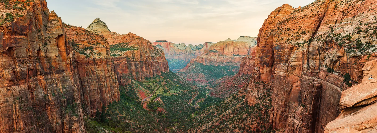 A wide panorama of red and orange canyon walls catching soft morning light, with green trees in the valley below.