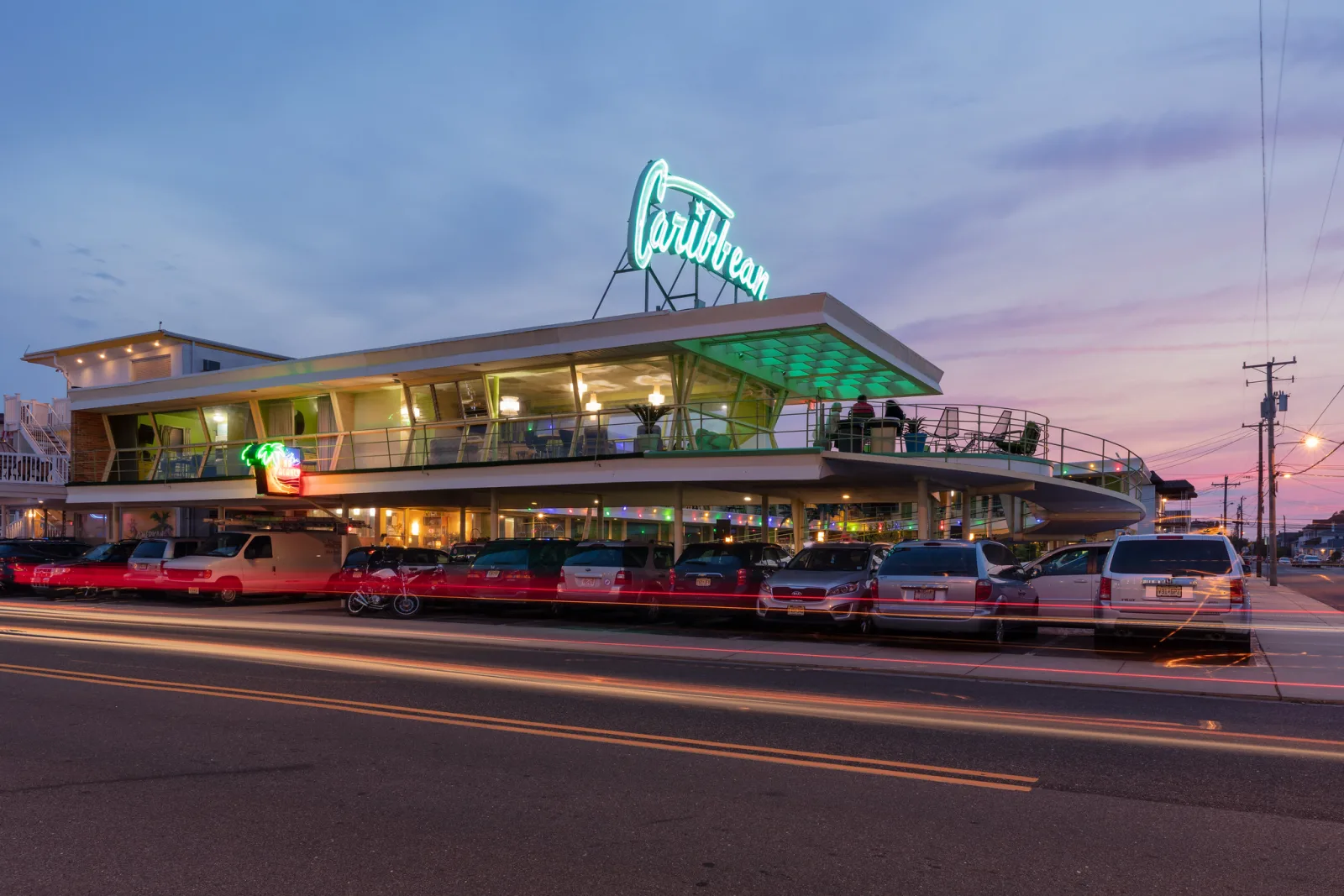 A two-storey motel with a lit green neon sign on its roof stands beside a street at dusk, with car light trails in front.