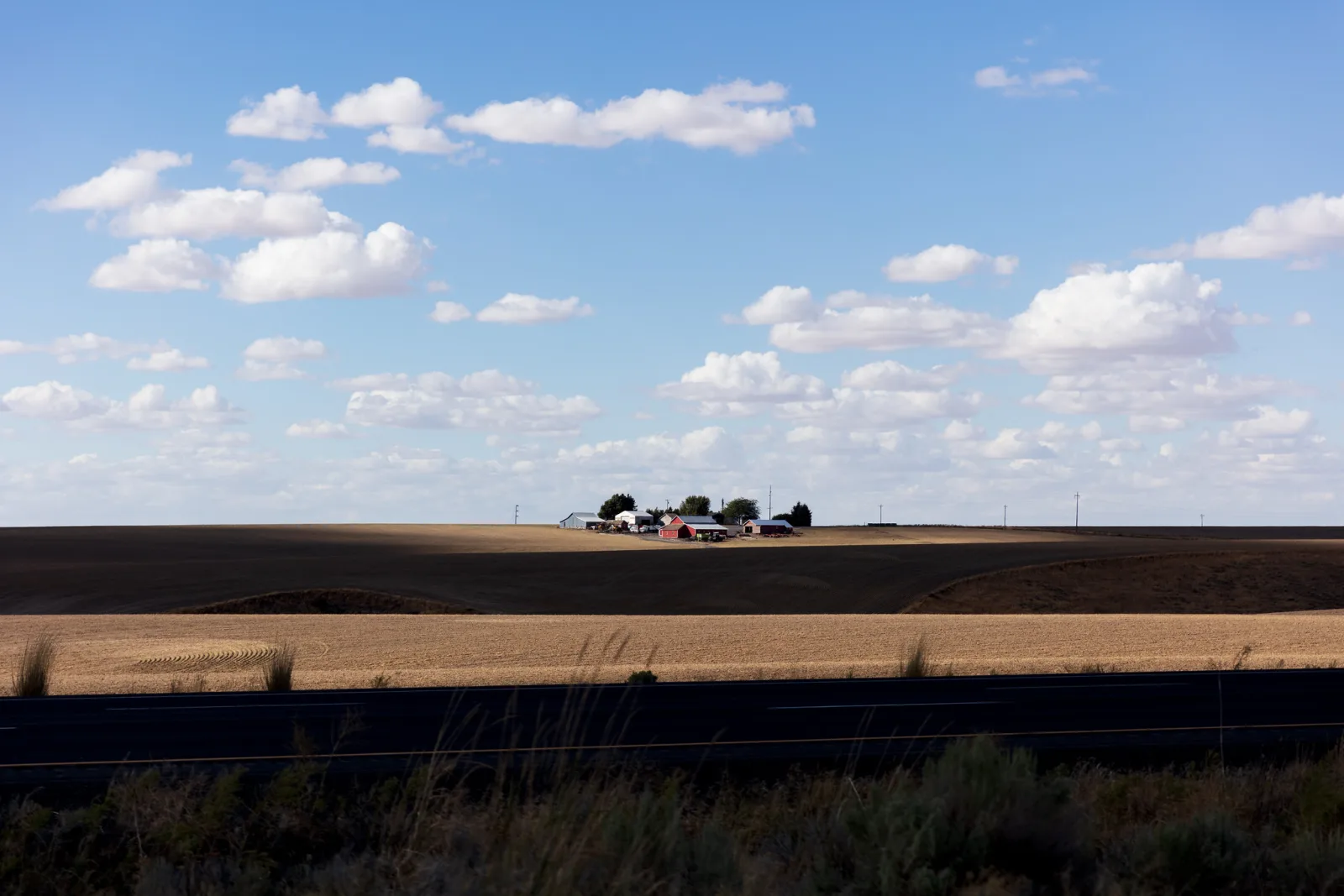 A farm sits in a wide plowed field under a blue sky with scattered clouds, with patches of shadow and sun across the ground.