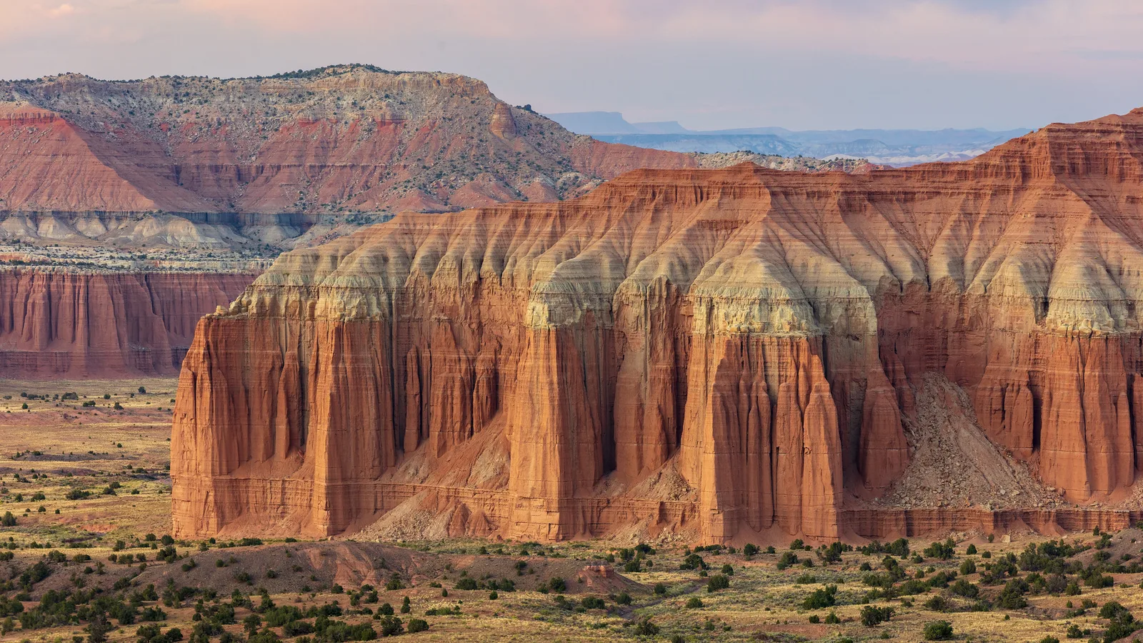 Tall red and cream sandstone cliffs rising from a desert valley in warm light
