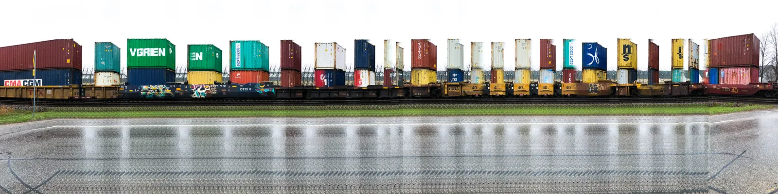 A long train of colourful shipping containers crosses the frame side-on, reflected in a strip of water in front of it.