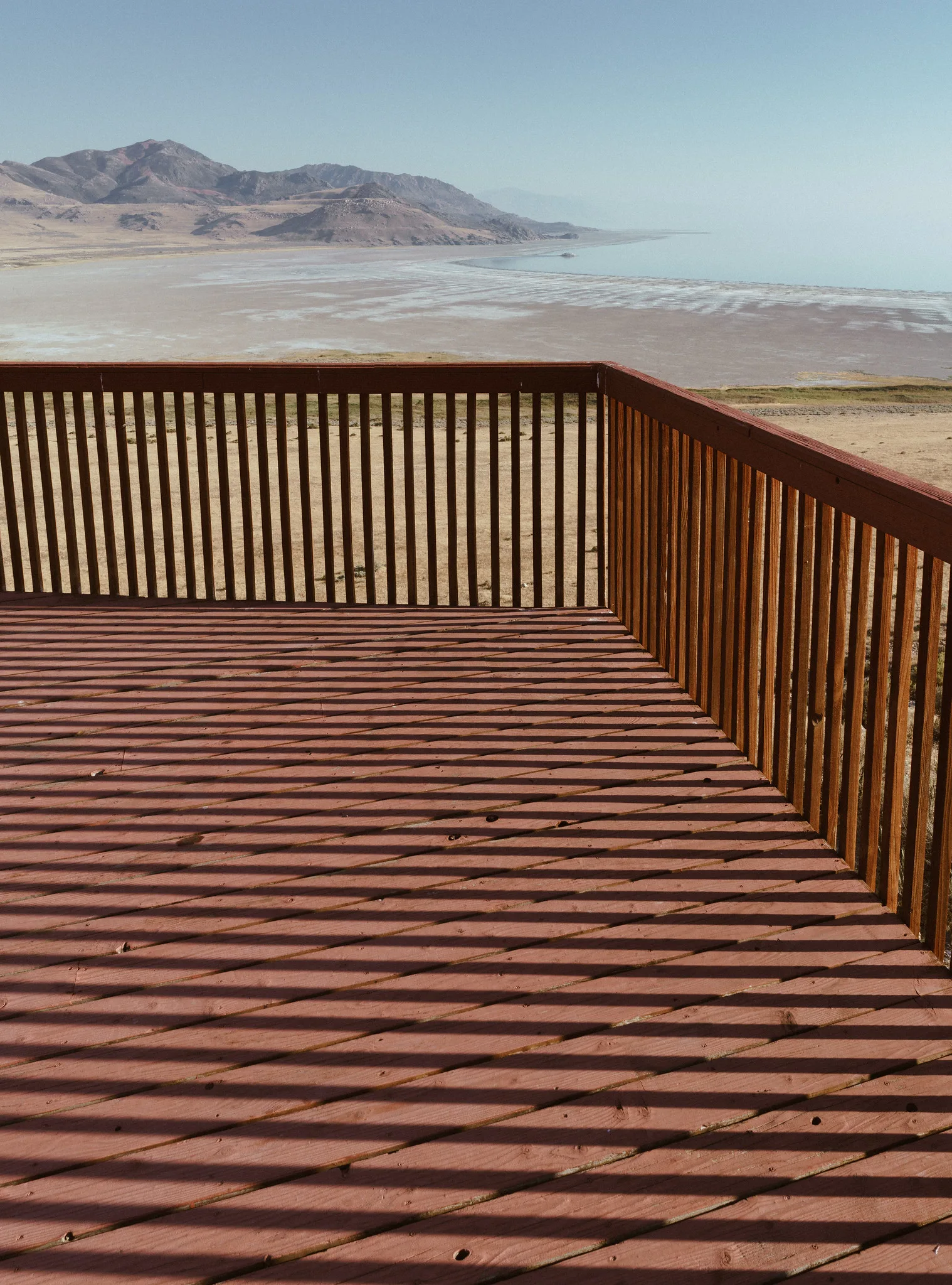 A wooden deck railing casts long bands of shadow across the red-brown deck boards in direct sun above a salt flat.