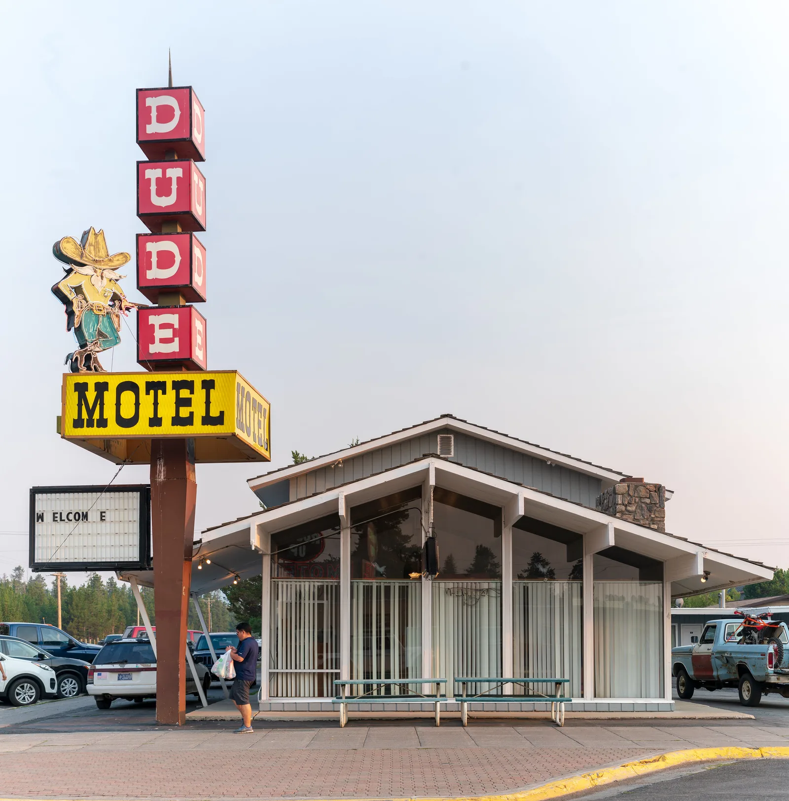 A tall vintage motel sign with red letter blocks, a cowboy figure and a yellow MOTEL panel stands beside a small motel office with large windows.