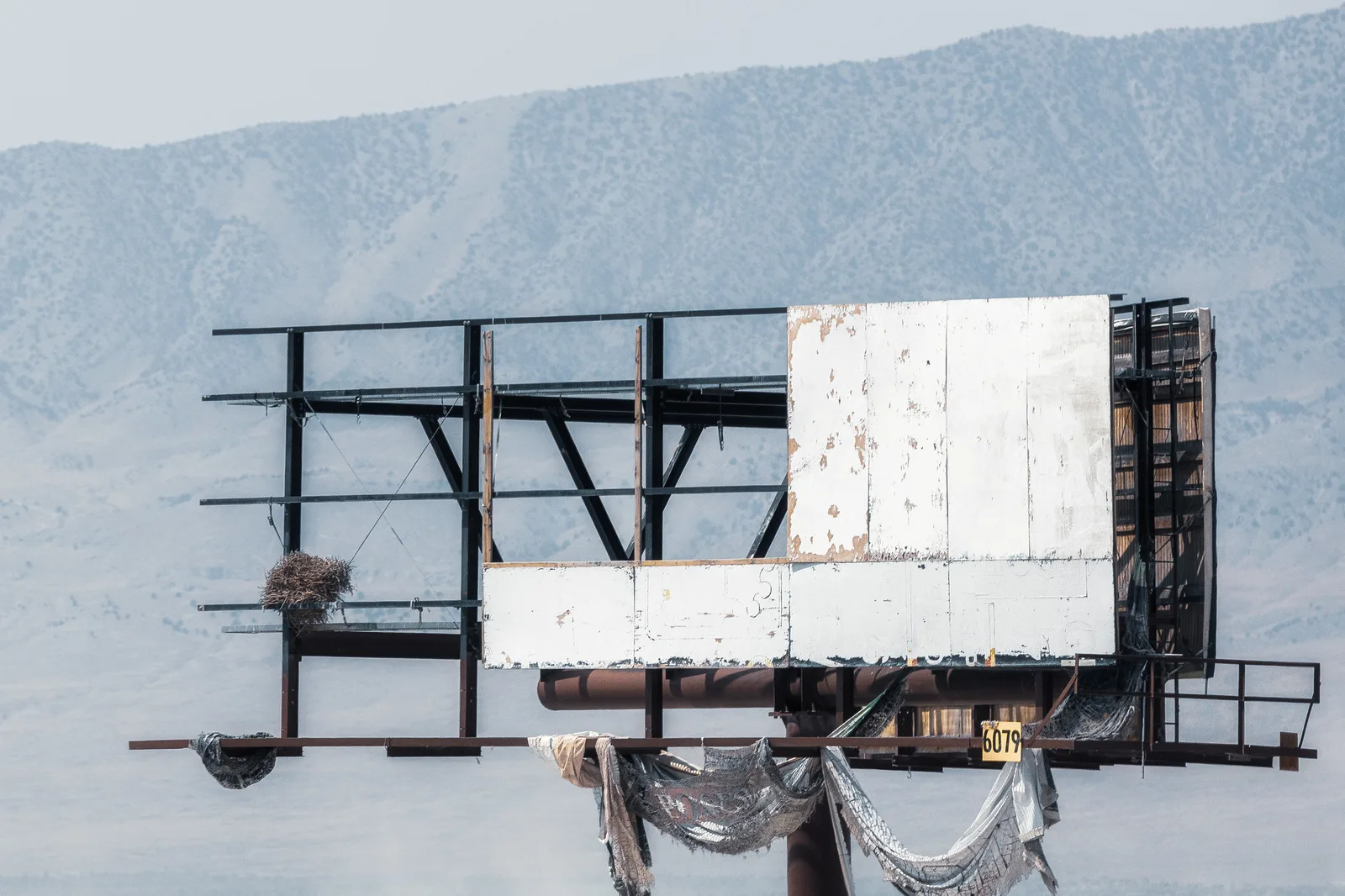 An empty, weathered billboard frame with peeling white panels and torn fabric hanging from its steel structure, in front of a hazy mountain slope.