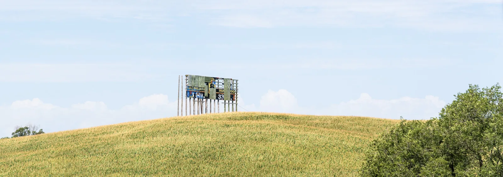 Peeling billboard on a rounded hill of pale green corn under a soft, washed-out blue sky with low clouds