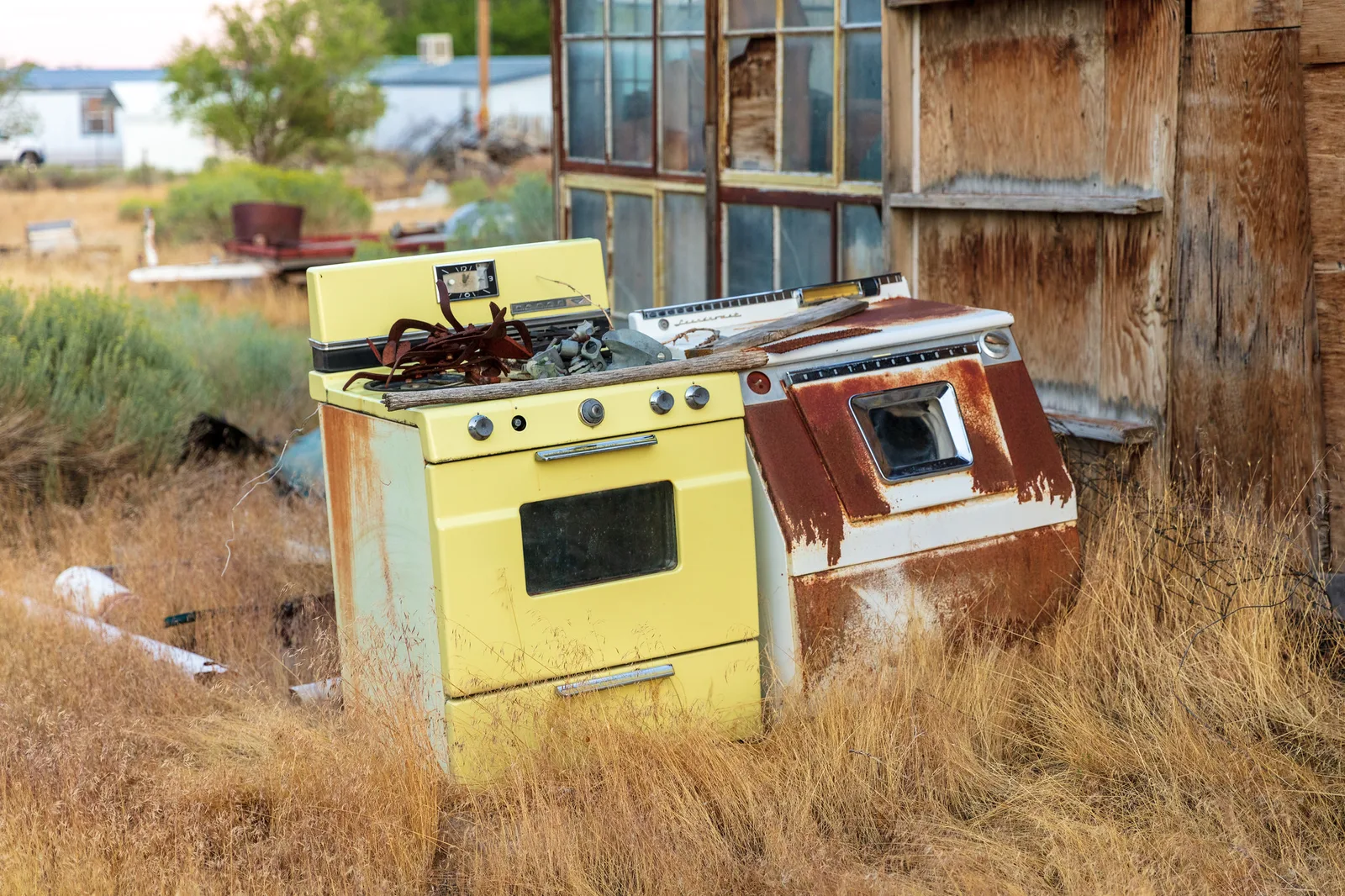 Two old kitchen stoves, one pale yellow and one streaked with rust, abandoned in dry golden grass beside a weathered wooden shed