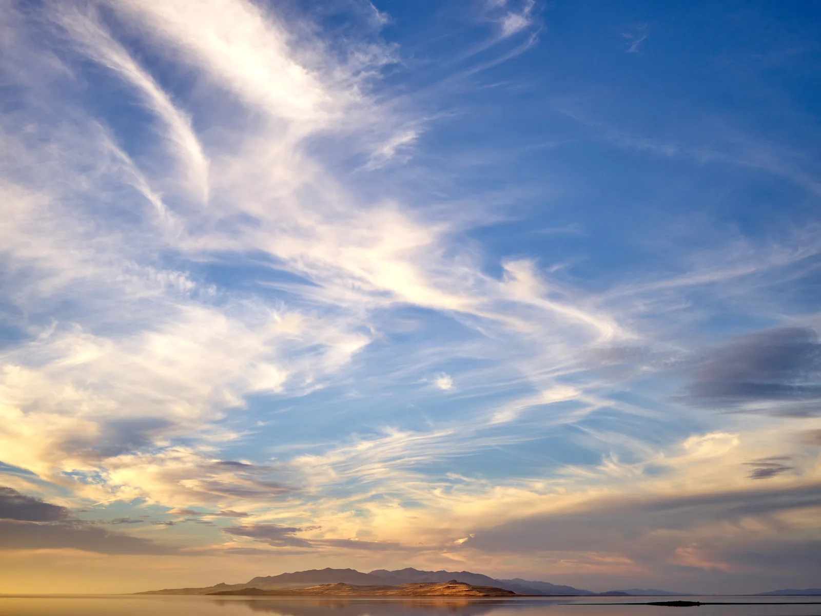 A wide sunset sky with streaky white and gold clouds over a lake, brightest near the setting sun