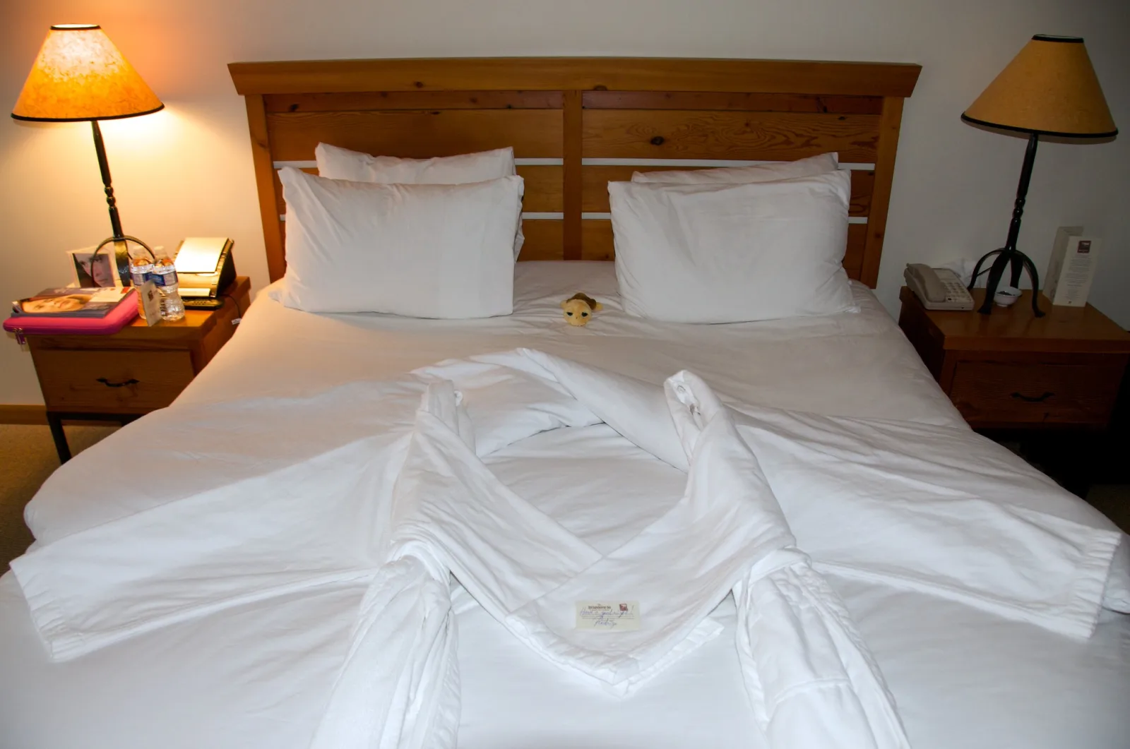 A made hotel bed with white linen, a wooden headboard and a lit lamp on each side.