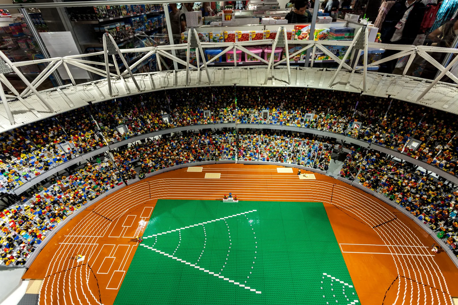 A model of an athletics stadium built from toy bricks, with stands full of tiny brick spectators around a green field and an orange track.