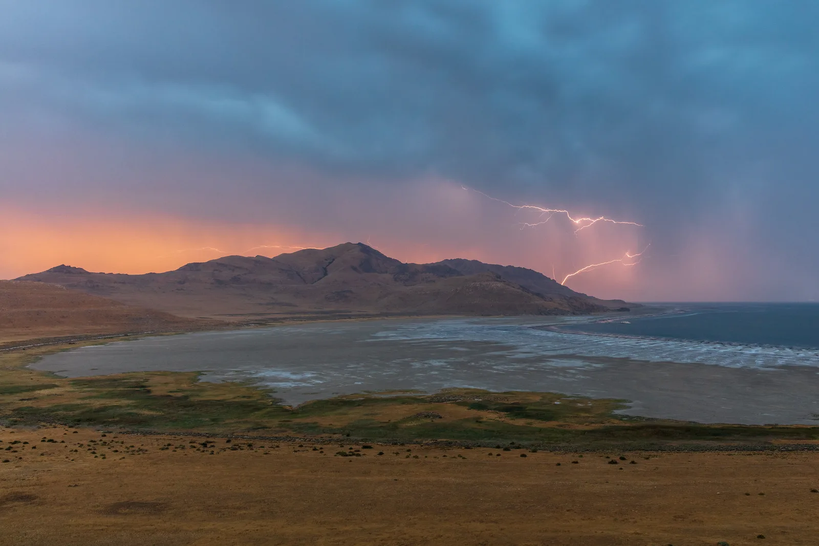 Lightning forks from a dark storm cloud over a shallow bay and a long ridge of hills, with a band of orange sunset light along the horizon.