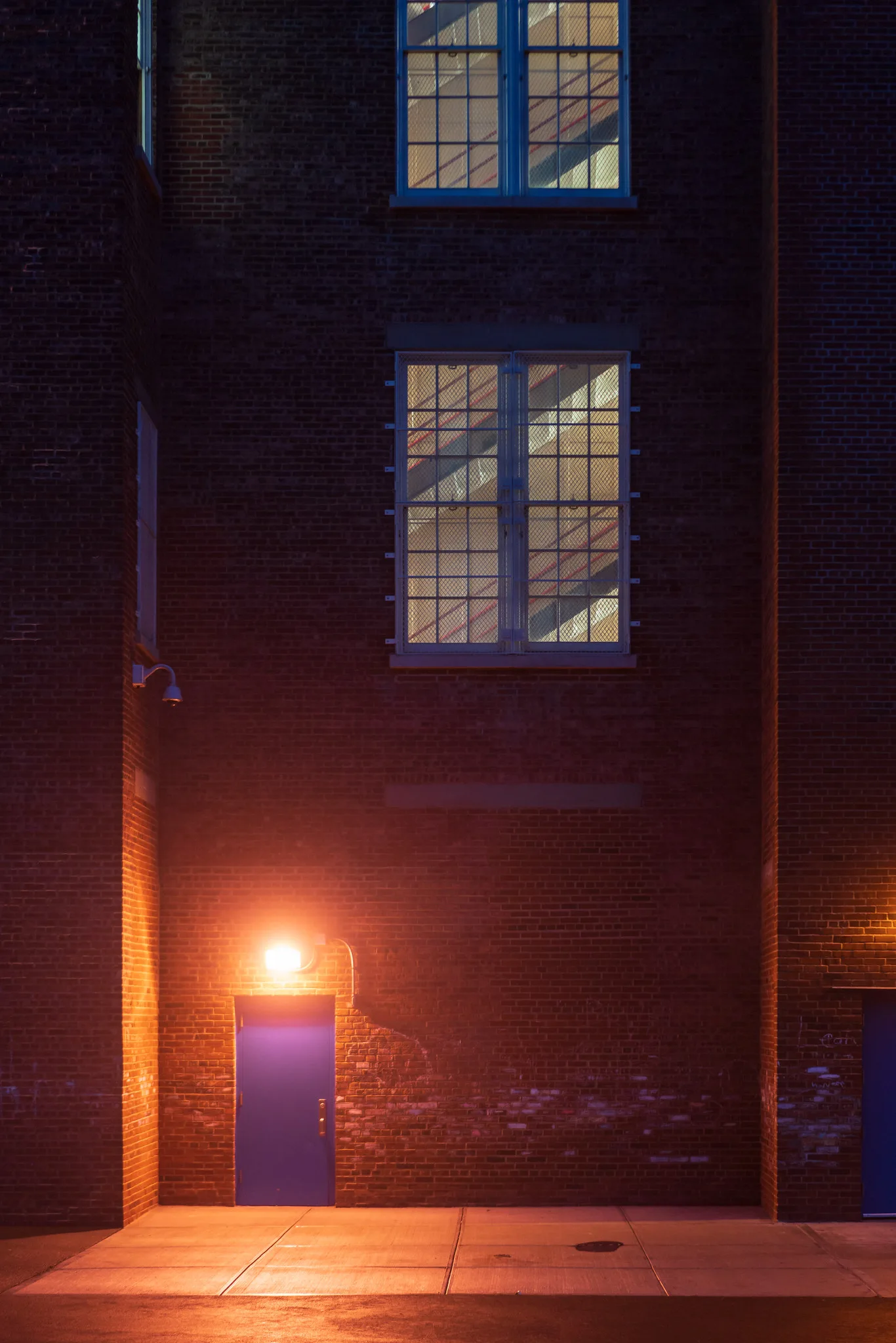 A blue door lit by a single warm lamp at the foot of a dark brick building at dusk, with tall lit windows above showing a staircase inside.