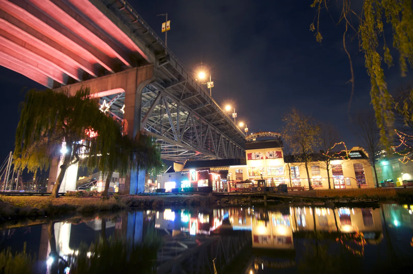 A bridge and a row of small lit buildings reflected in water at night, with street lamps glowing in the dark.