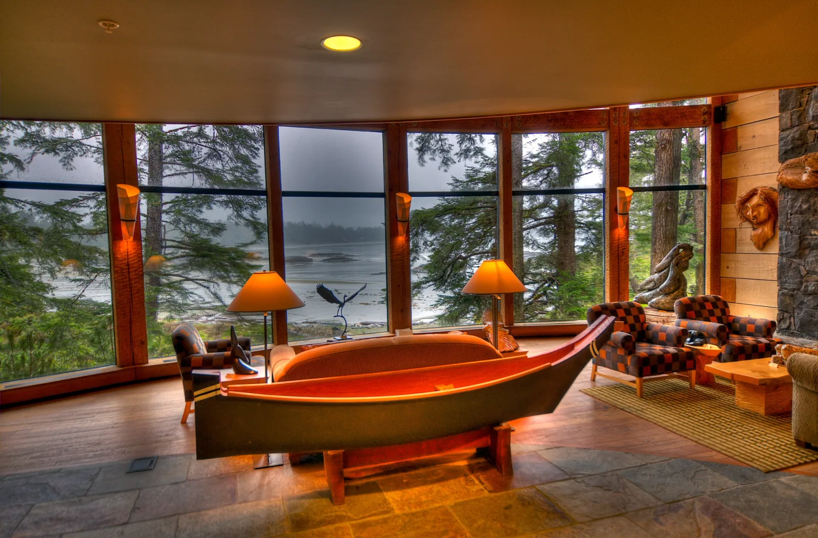A lodge lounge with warm lamps lit beside large windows that look out on trees and a grey shoreline.