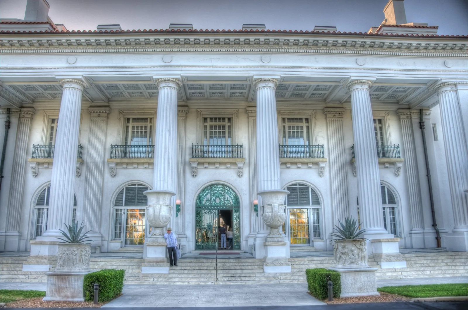 The white marble front of a grand mansion museum with tall columns, a balcony and arched doorways behind a trimmed lawn.