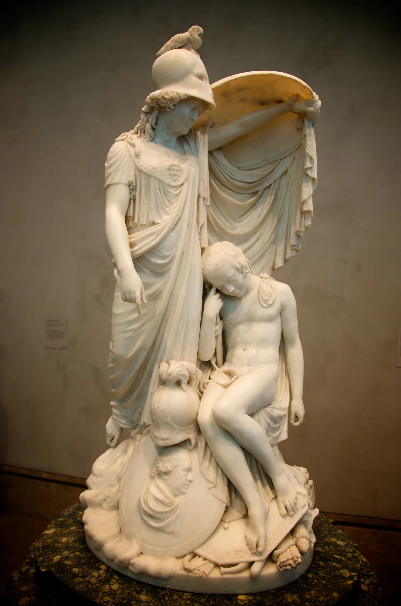 A white marble sculpture of a standing robed figure with a smaller figure at her side, lit warmly against a dark background.