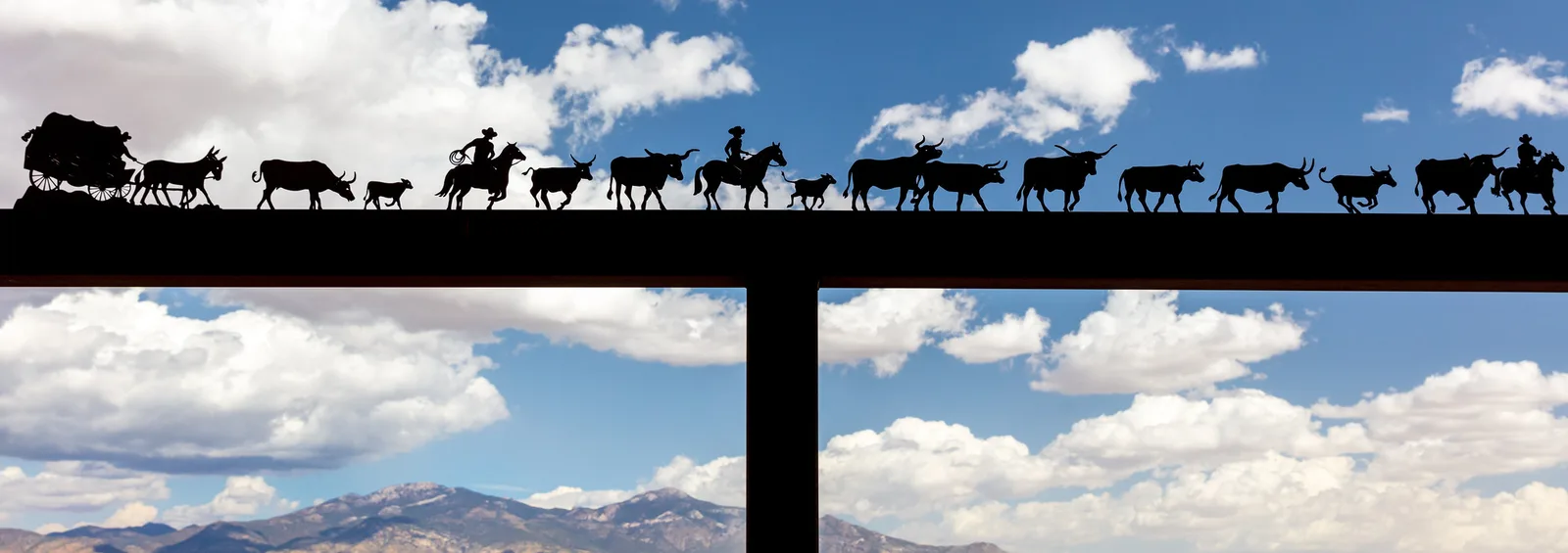 A dark metal sculpture of cowboys driving cattle across a beam, silhouetted against a bright blue sky with white clouds