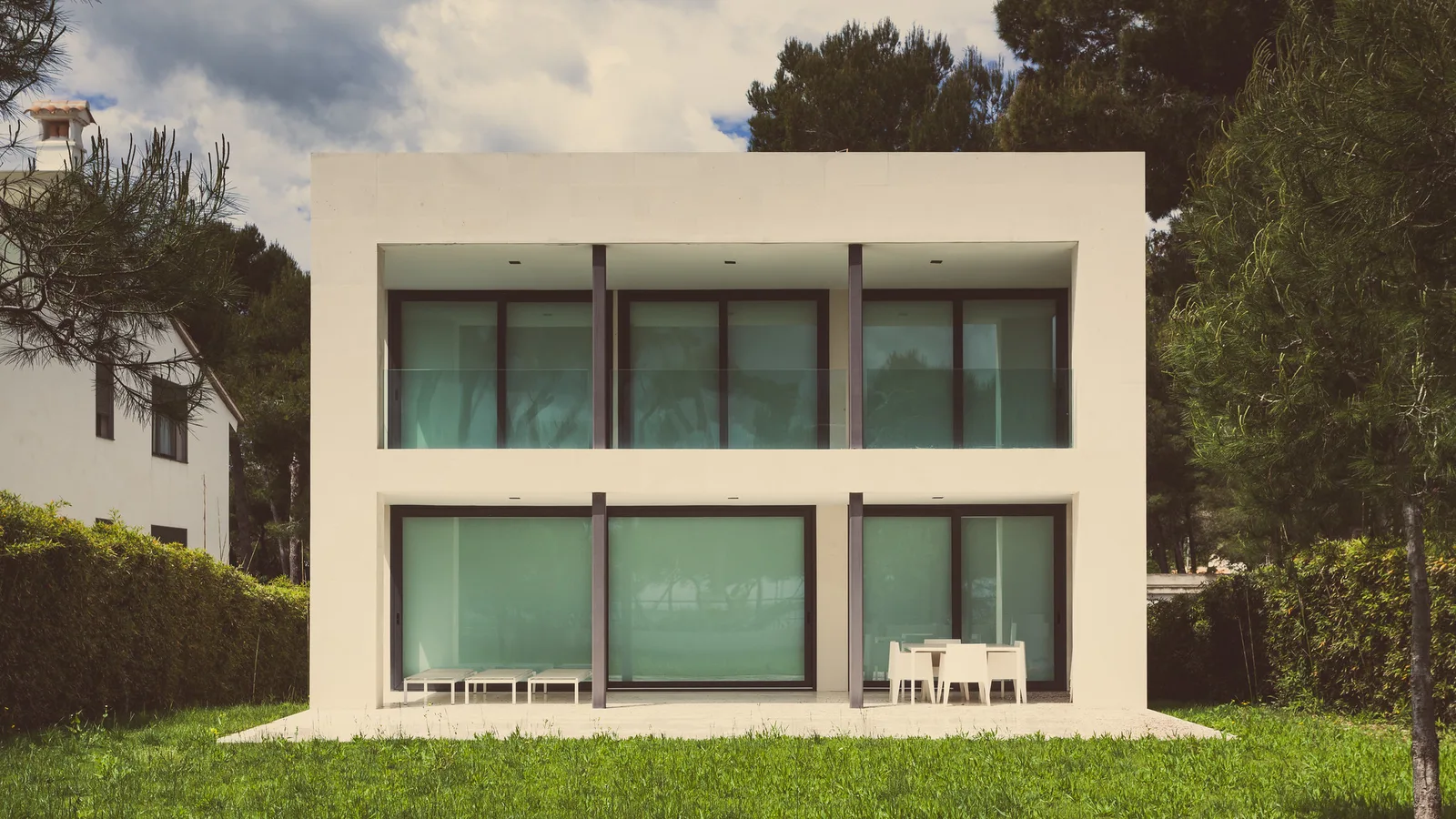 A modern two-story white house with large dark-framed windows and a terrace, set on a lawn between tall pine trees.