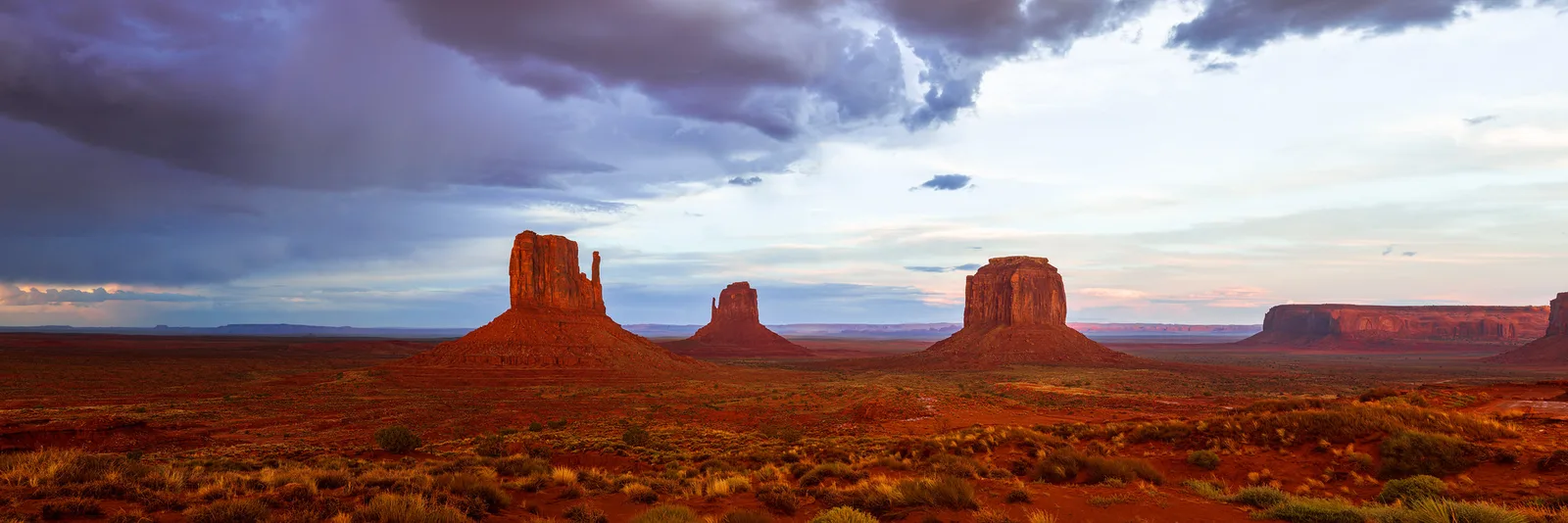 Red sandstone buttes stand in a wide desert valley glowing in late light as dark rain clouds clear overhead.