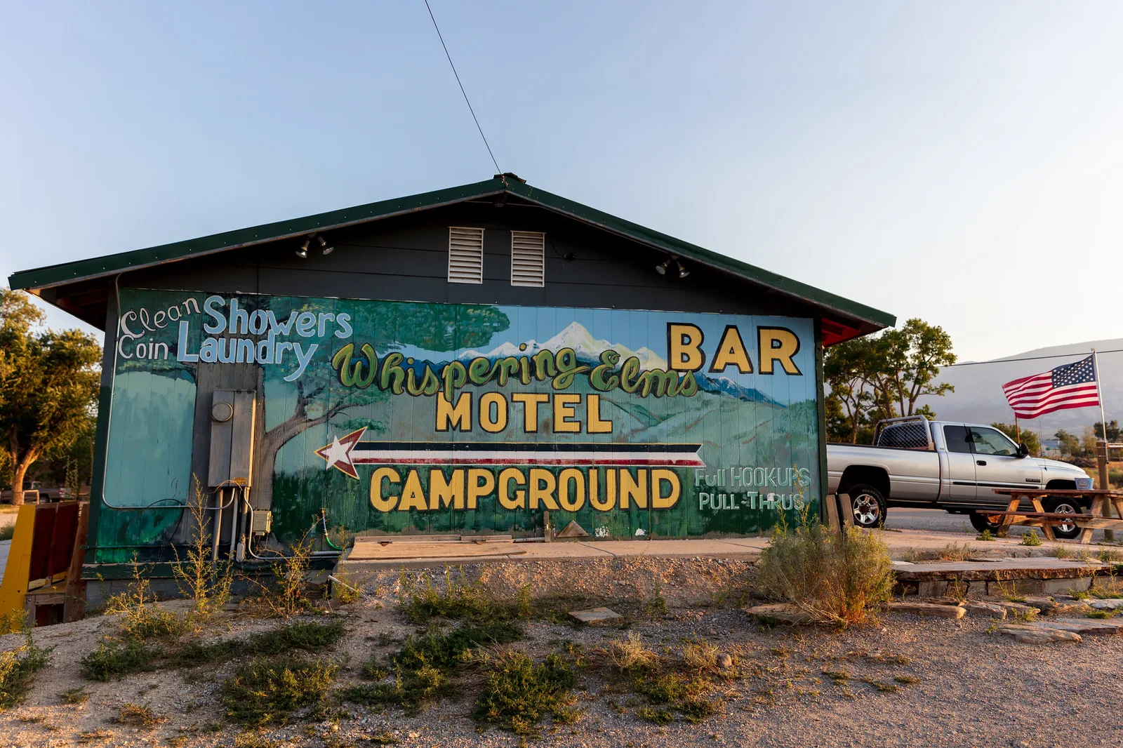 A roadside building painted with a large mural advertising a bar, motel and campground, with a pickup truck parked beside it.