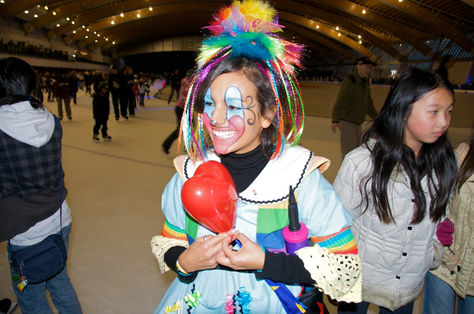 A face-painted clown performer with a rainbow wig grinning at a crowded indoor skating event