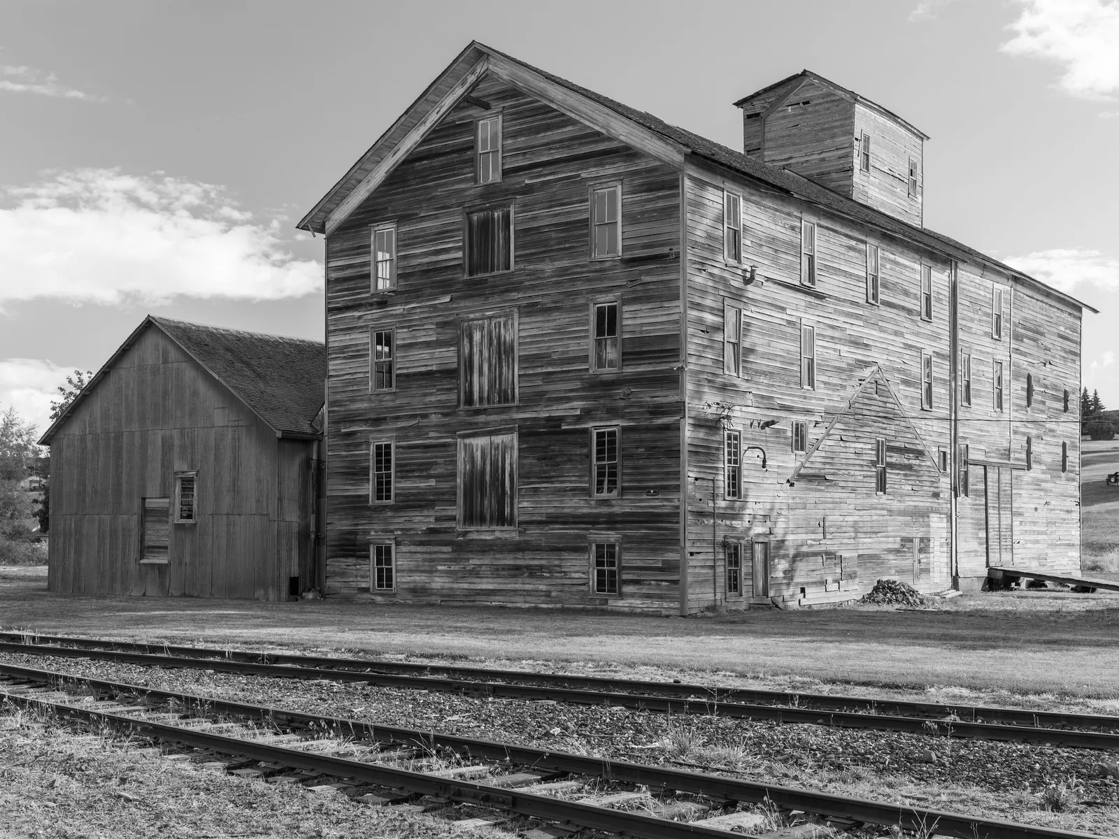 Black and white photo of a tall weathered wooden mill building and a smaller shed beside railway tracks under a cloudy sky.
