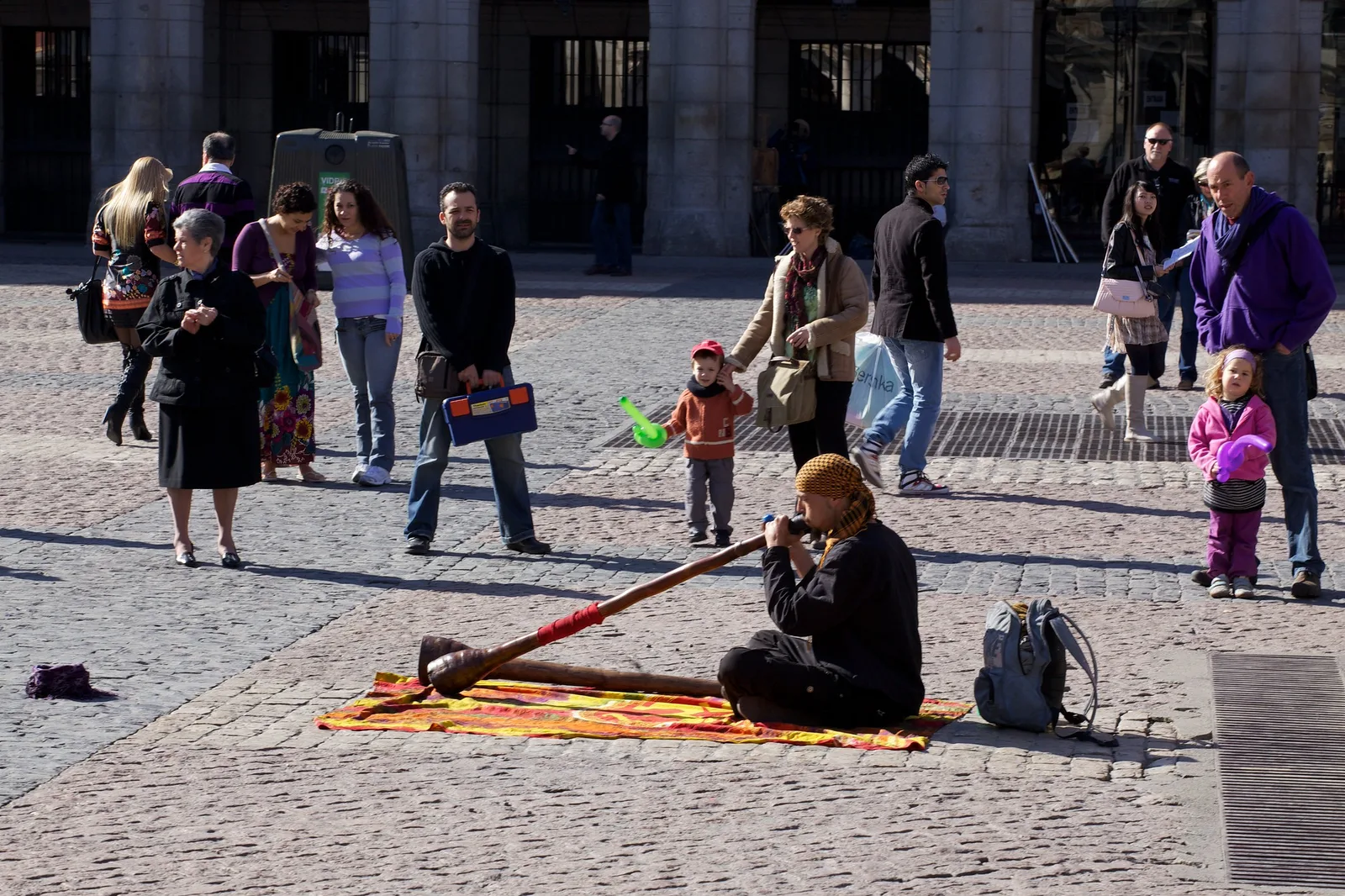 A street musician seated on a mat in a Madrid square, both hands holding a long didgeridoo
