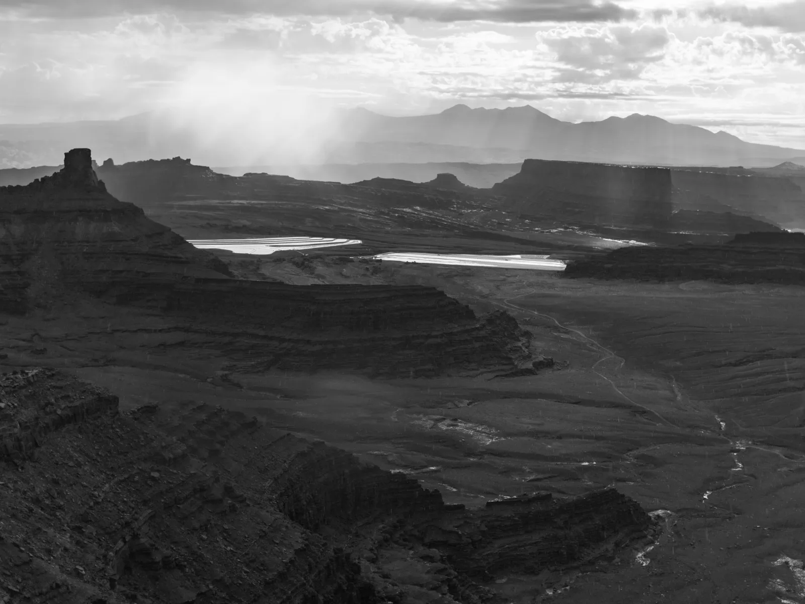 A black and white view over layered canyon land with bright cloud and rain above dark rock.
