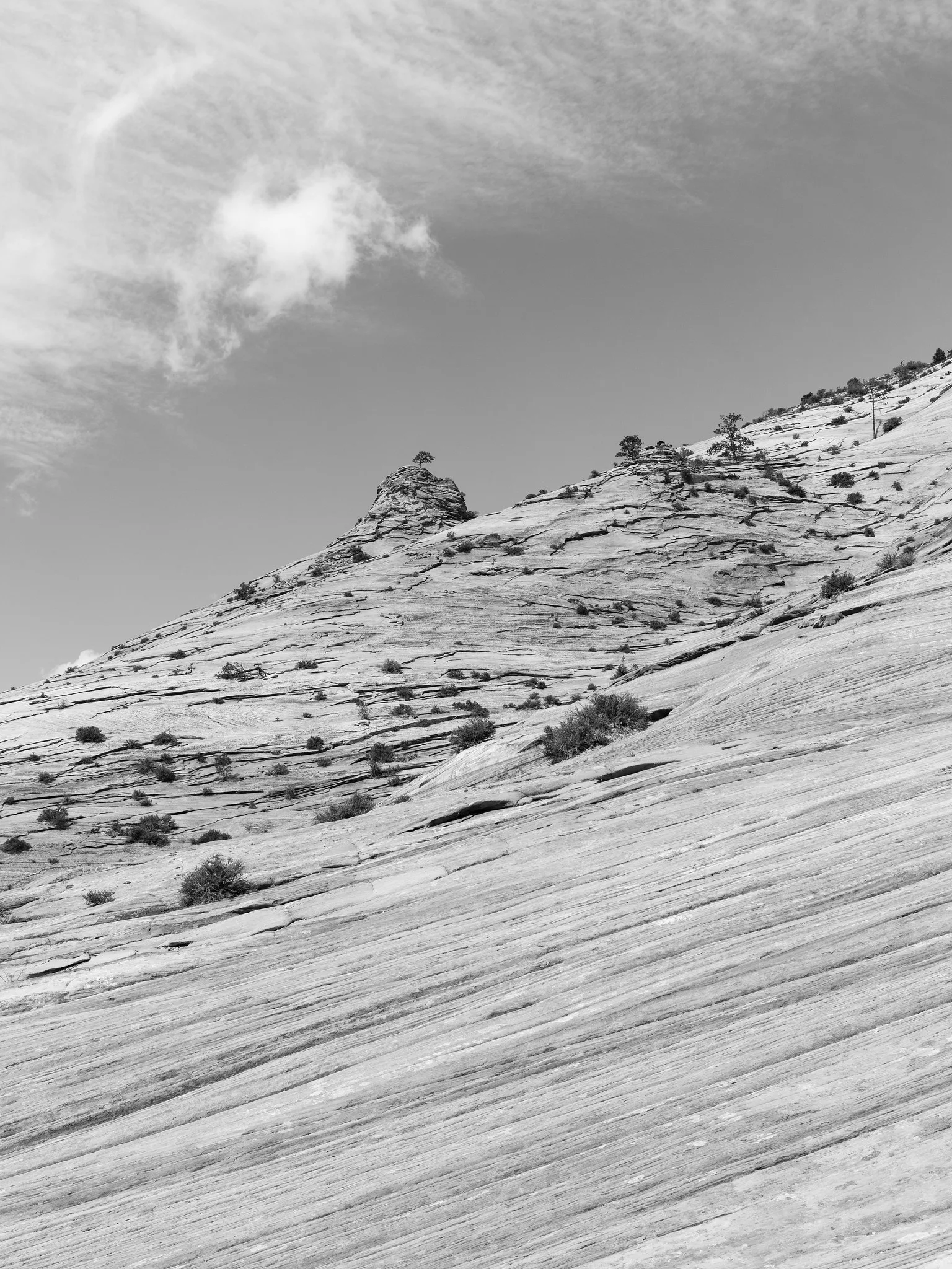Black and white photo of a smooth sandstone slope with fine curving lines rising to rocky outcrops under a cloudy sky.